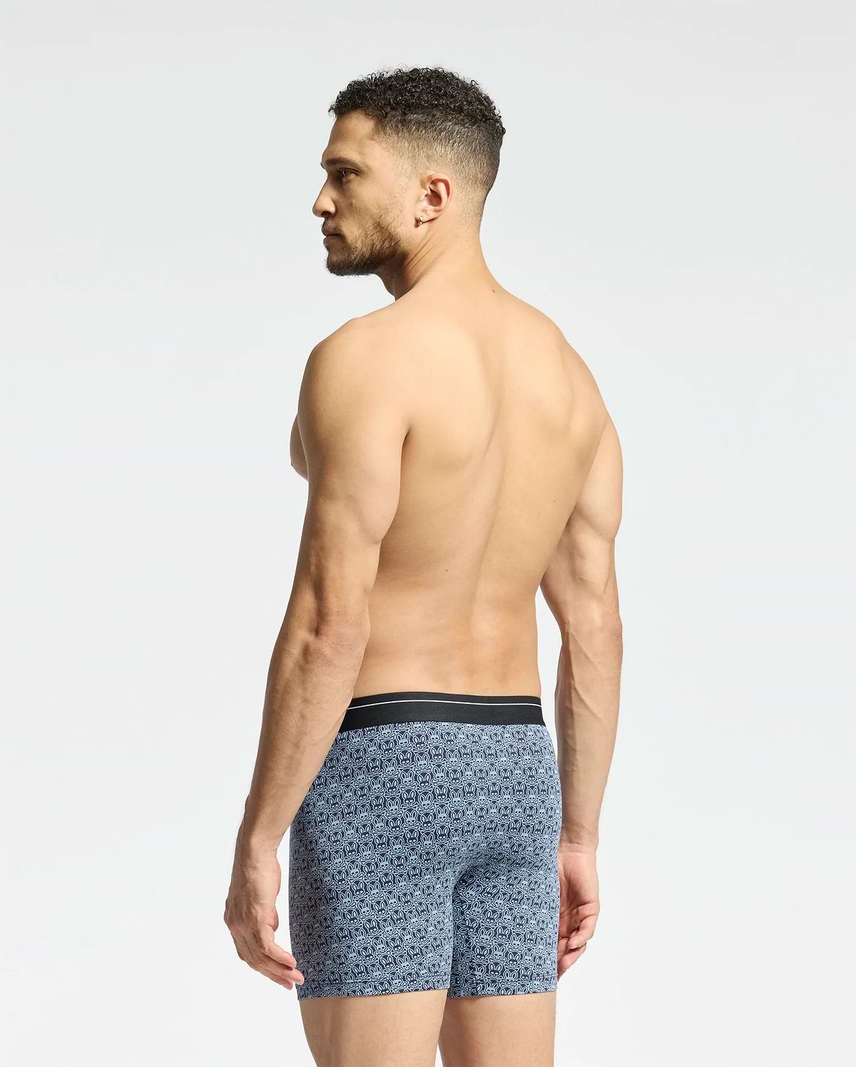 A man stands with his back turned, wearing Psycho Bunny's MENS 2 PACK BOXER BRIEF - B6V481F200 crafted from stretch Pima cotton in a blue pattern against a plain light gray background.