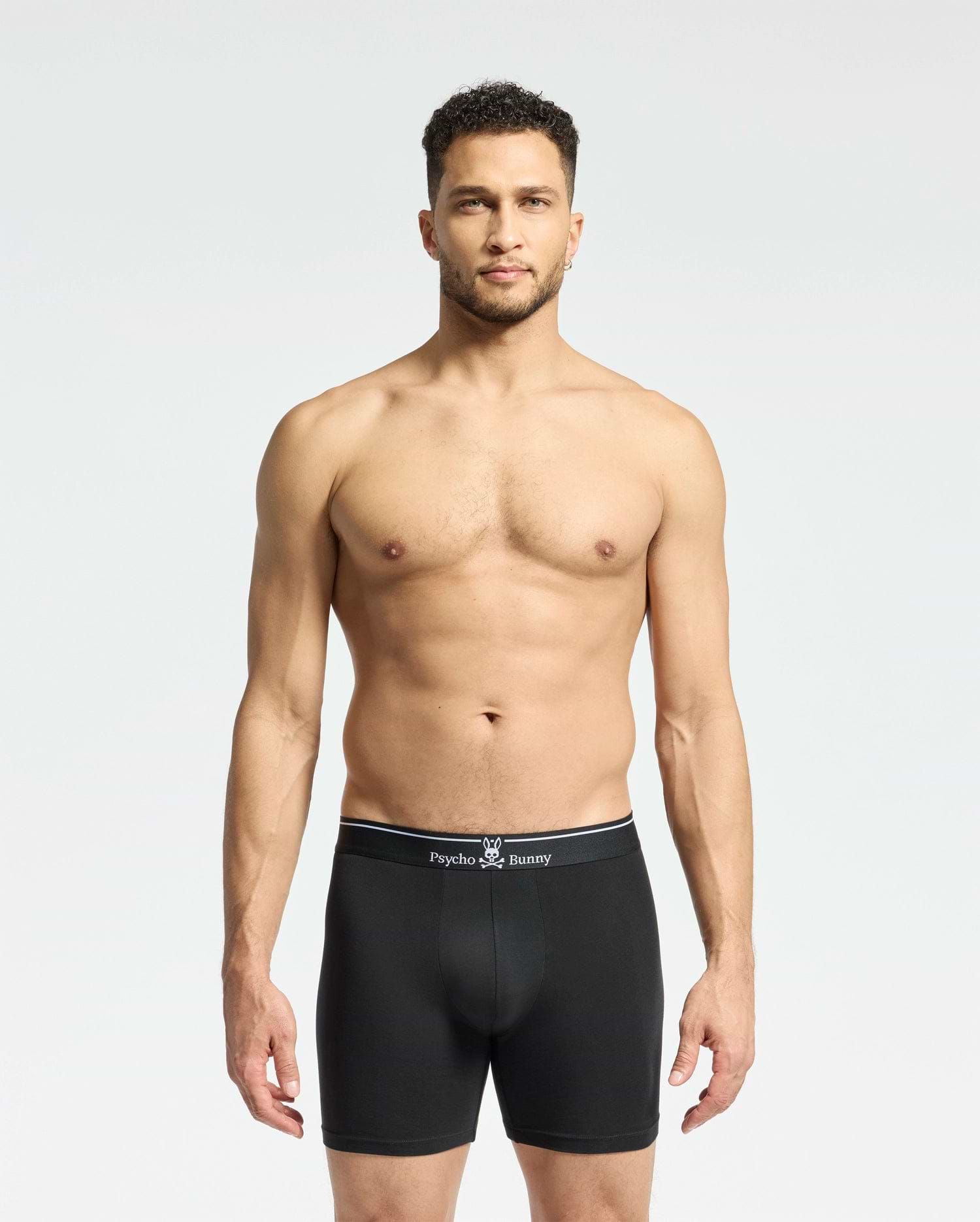 A man with short curly hair faces forward, wearing Psycho Bunny MENS SOLID KNIT 2 PACK BOXER BRIEF (B6V906ARCM) with branded waistband, made from soft Pima cotton, against a plain light background.