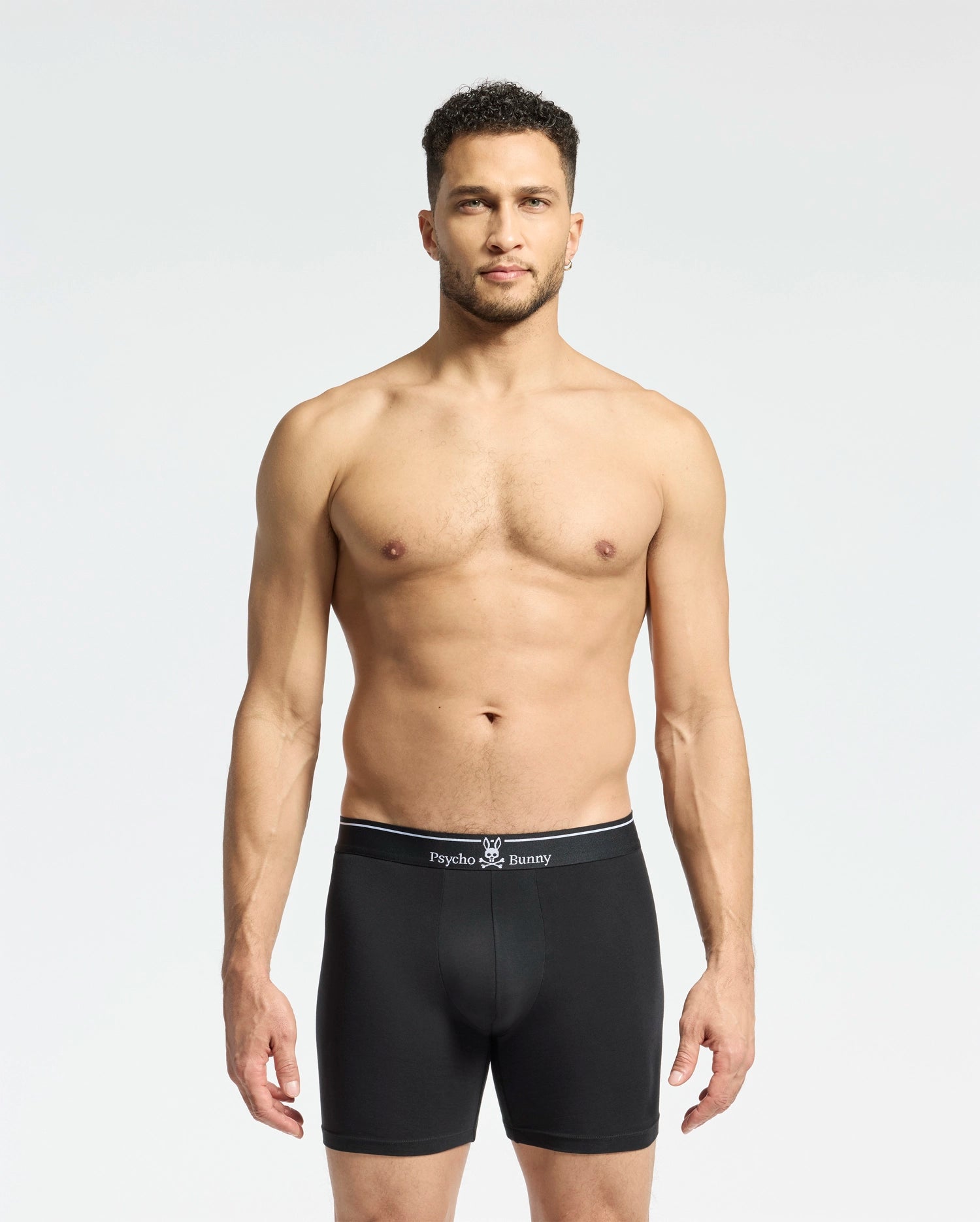 A man with short curly hair faces forward, wearing Psycho Bunny MENS SOLID KNIT 2 PACK BOXER BRIEF (B6V906ARCM) with branded waistband, made from soft Pima cotton, against a plain light background.