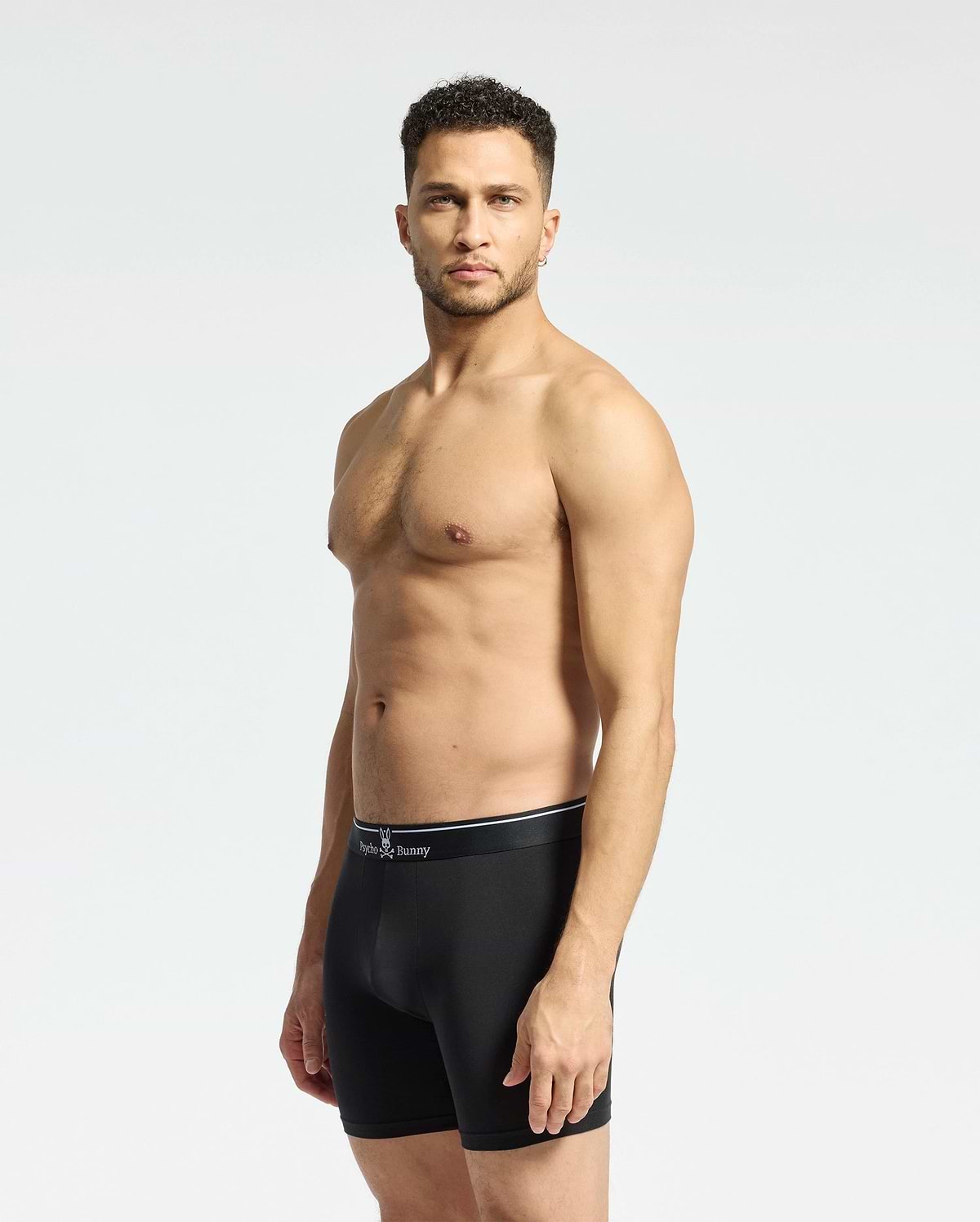 A man with short curly hair stands against a plain light backdrop, wearing Psycho Bunny MENS SOLID KNIT 2 PACK BOXER BRIEF - B6V906ARCM in black. He faces slightly to the side with a neutral expression.