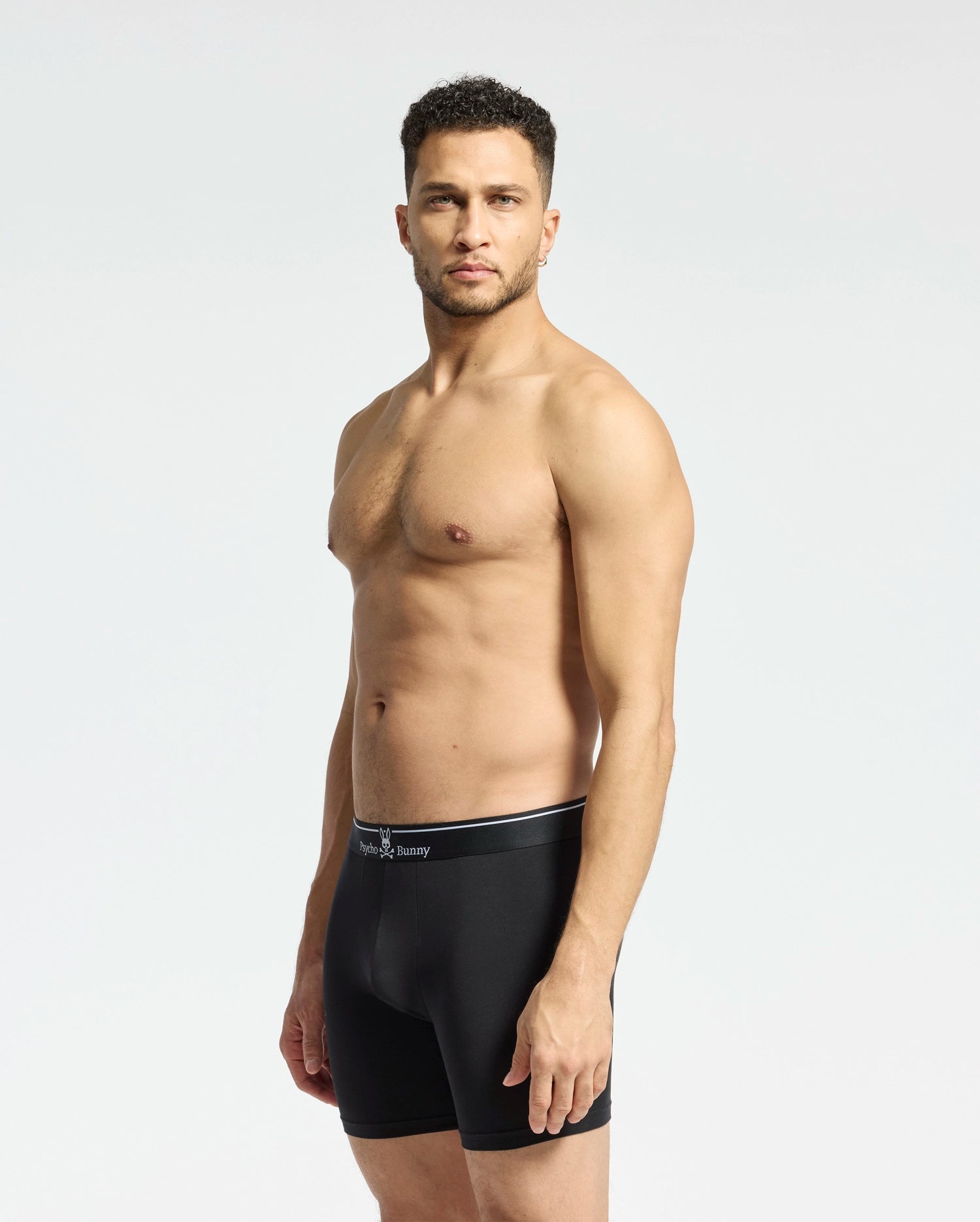 A man with short curly hair stands against a plain light backdrop, wearing Psycho Bunny MENS SOLID KNIT 2 PACK BOXER BRIEF - B6V906ARCM in black. He faces slightly to the side with a neutral expression.