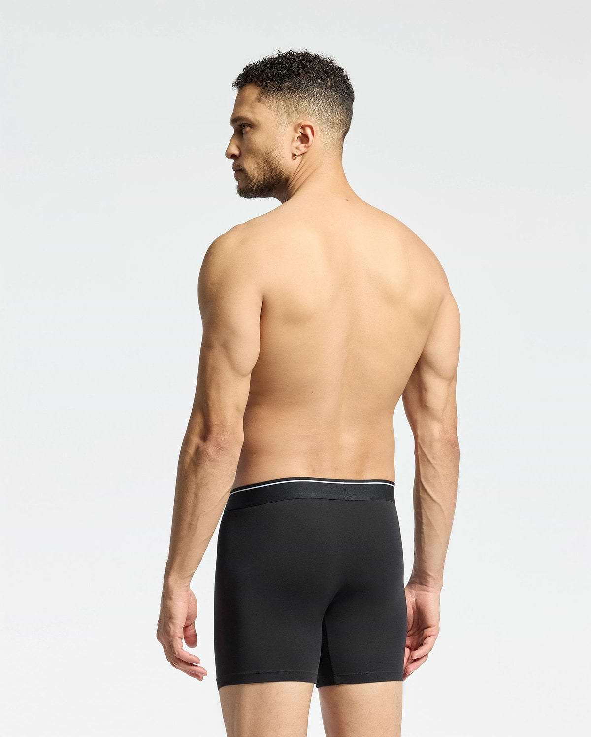 A man with short curly hair stands shirtless, facing away in Psycho Bunny MENS SOLID KNIT 2 PACK BOXER BRIEF (B6V906ARCM), black, against a plain light background.