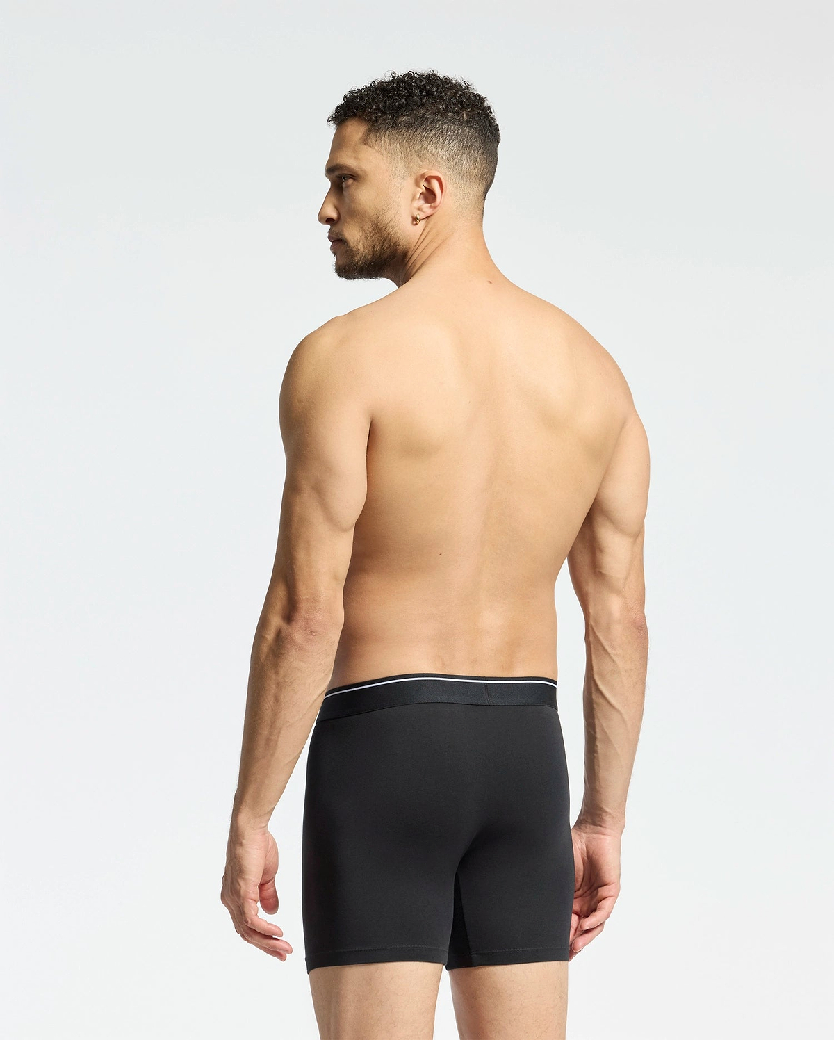 A man with short curly hair stands shirtless, facing away in Psycho Bunny MENS SOLID KNIT 2 PACK BOXER BRIEF (B6V906ARCM), black, against a plain light background.