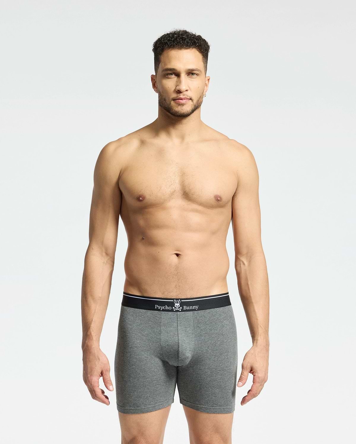 A man stands against a plain background, wearing gray fitted boxers from the Psycho Bunny MENS SOLID KNIT 2 PACK. These Pima cotton briefs feature a black waistband with the brand's bunny logo and crossbones, epitomizing style and comfort.