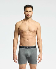 A man stands against a plain background, wearing gray fitted boxers from the Psycho Bunny MENS SOLID KNIT 2 PACK. These Pima cotton briefs feature a black waistband with the brand's bunny logo and crossbones, epitomizing style and comfort.