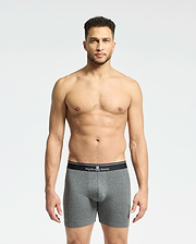 A man stands against a plain background, wearing gray fitted boxers from the Psycho Bunny MENS SOLID KNIT 2 PACK. These Pima cotton briefs feature a black waistband with the brand's bunny logo and crossbones, epitomizing style and comfort.