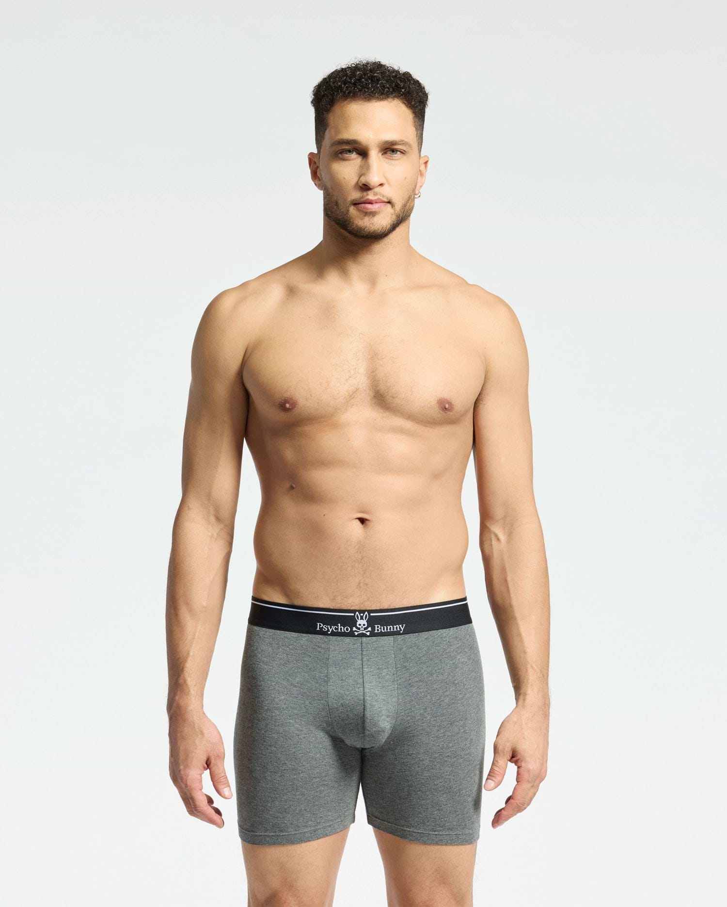 A man stands against a plain background, wearing gray fitted boxers from the Psycho Bunny MENS SOLID KNIT 2 PACK. These Pima cotton briefs feature a black waistband with the brand's bunny logo and crossbones, epitomizing style and comfort.