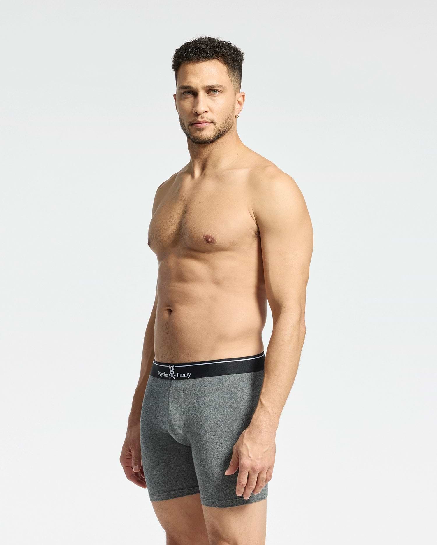 A man with short curly hair stands against a plain background, wearing Psycho Bunny's MENS SOLID KNIT 2 PACK BOXER BRIEF - B6V906ARCM. The gray fitted boxer briefs, crafted from soft Pima cotton with a black waistband, offer comfort as he maintains a neutral expression.