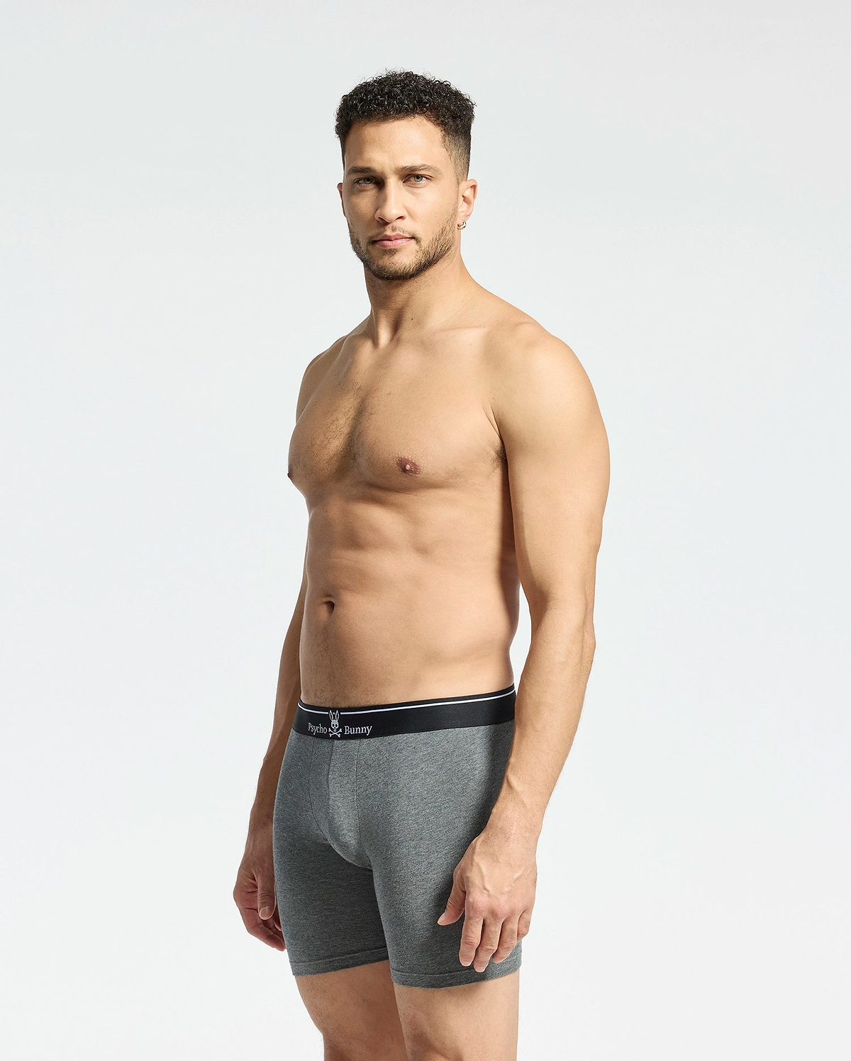 A man with short curly hair stands against a plain background, wearing Psycho Bunny's MENS SOLID KNIT 2 PACK BOXER BRIEF - B6V906ARCM. The gray fitted boxer briefs, crafted from soft Pima cotton with a black waistband, offer comfort as he maintains a neutral expression.