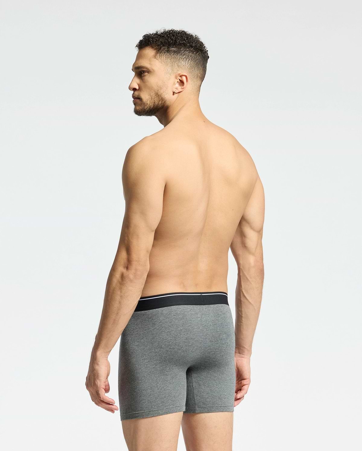 A shirtless man with short curly hair faces away, displaying Psycho Bunny's MENS SOLID KNIT 2 PACK BOXER BRIEF - B6V906ARCM in gray. These comfy Pima cotton boxers feature a sleek black waistband. He stands against a plain white background.