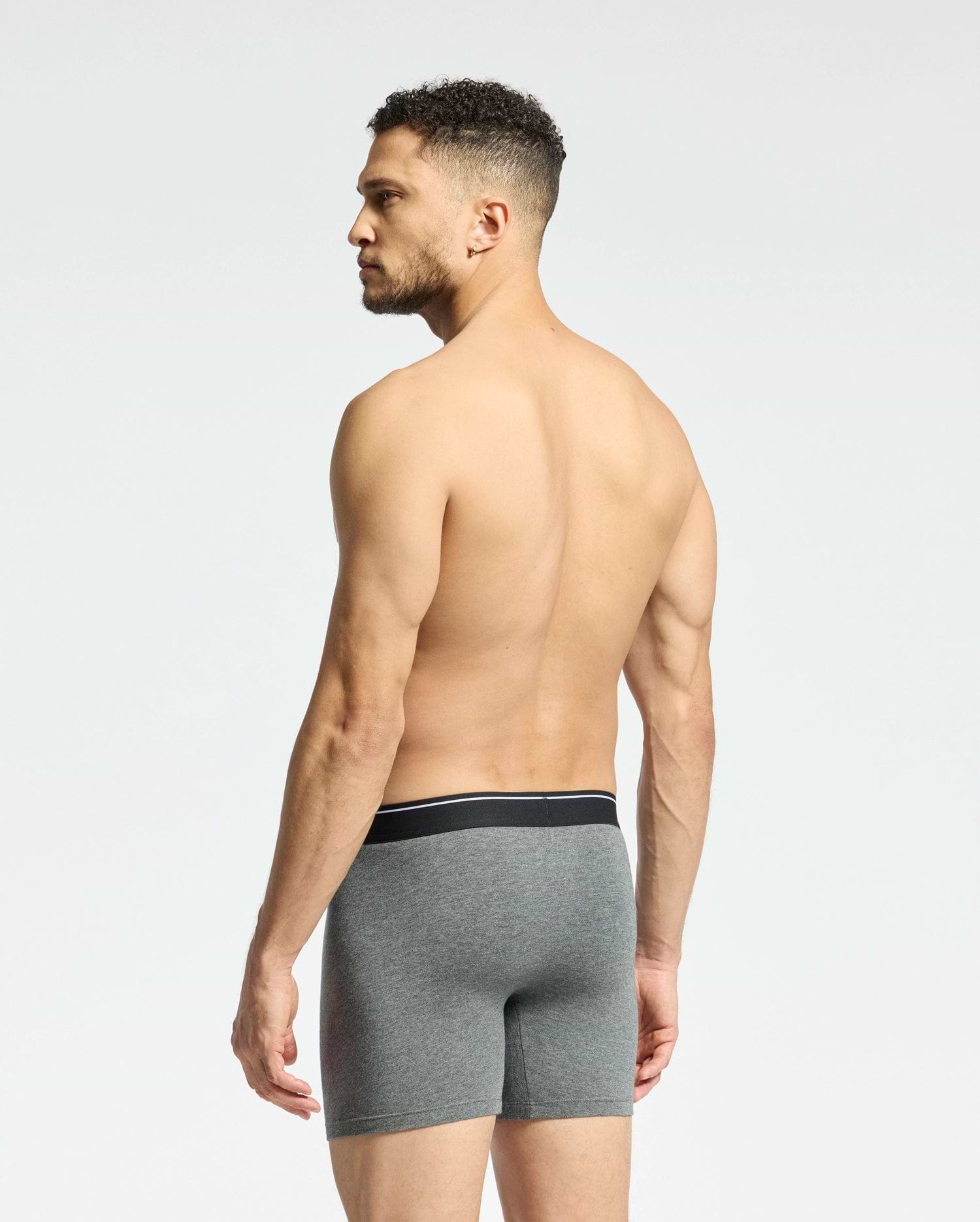 A shirtless man with short curly hair faces away, displaying Psycho Bunny's MENS SOLID KNIT 2 PACK BOXER BRIEF - B6V906ARCM in gray. These comfy Pima cotton boxers feature a sleek black waistband. He stands against a plain white background.