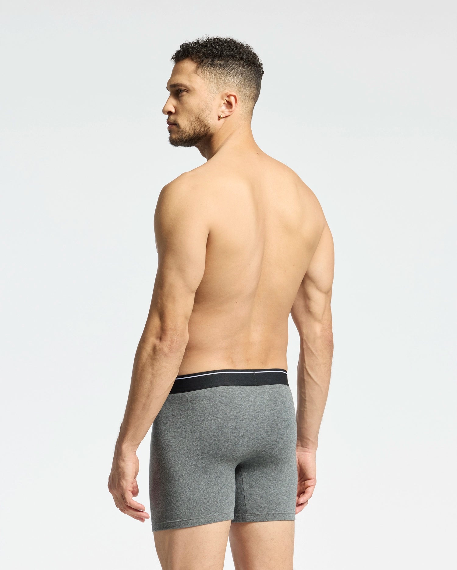 A shirtless man with short curly hair faces away, displaying Psycho Bunny's MENS SOLID KNIT 2 PACK BOXER BRIEF - B6V906ARCM in gray. These comfy Pima cotton boxers feature a sleek black waistband. He stands against a plain white background.