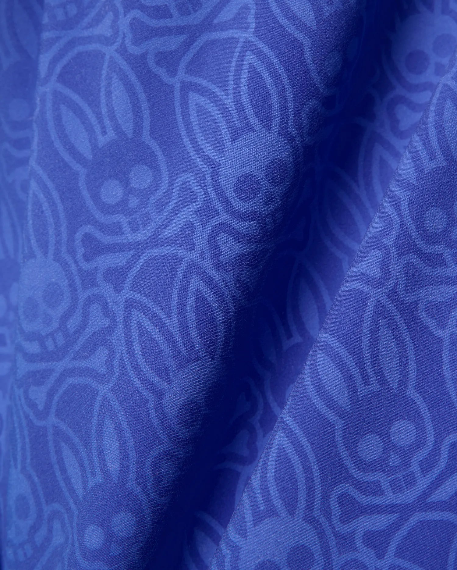 Mens Samos Hydrochromic Printed Swim Trunk in royal blue, featuring a subtle tonal all-over Psycho Bunny skull and crossbones logo pattern on smooth fabric.