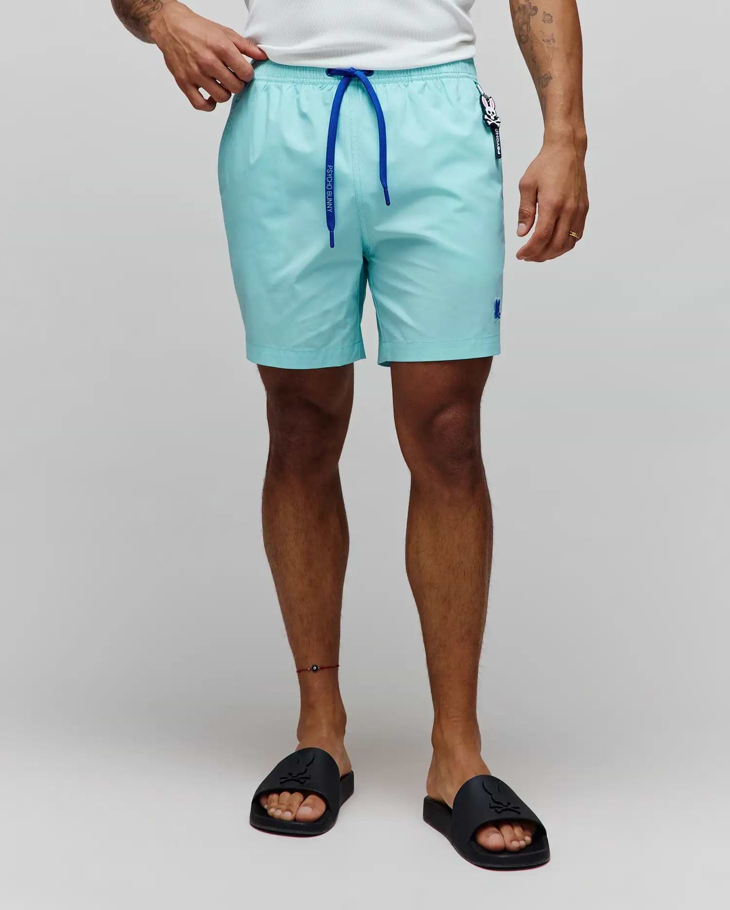 Mens Samos Hydrochromic Printed Swim Trunk in aqua, featuring a smooth texture, royal blue drawstring with Psycho Bunny text, small embroidered blue bunny skull logo on the left leg, and a branded tag on the right hip.