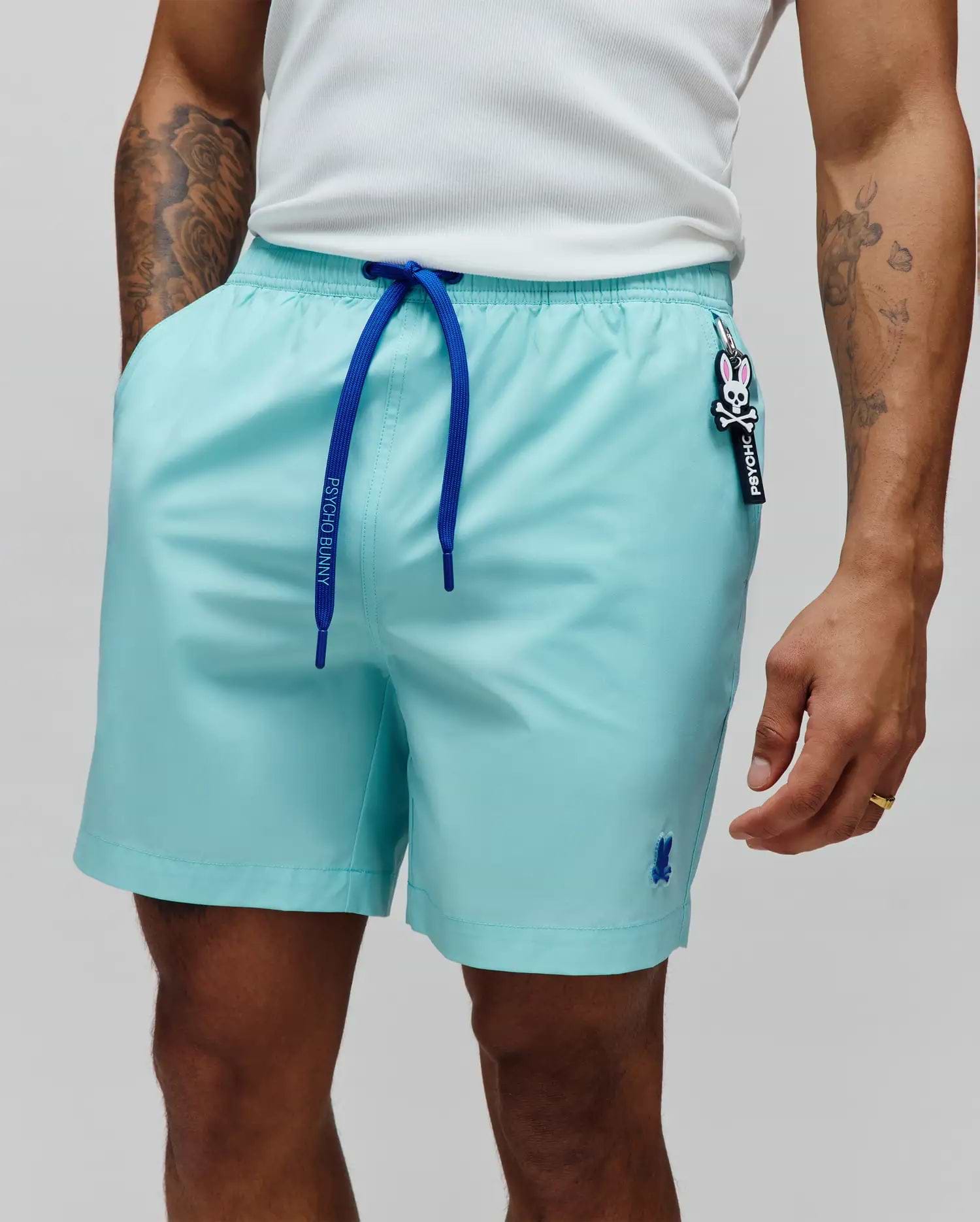 Mens Samos Hydrochromic Printed Swim Trunk, relaxed aqua with royal blue Psycho Bunny drawstrings, small embroidered blue bunny skull logo on left leg, and a Psycho Bunny skull keychain.