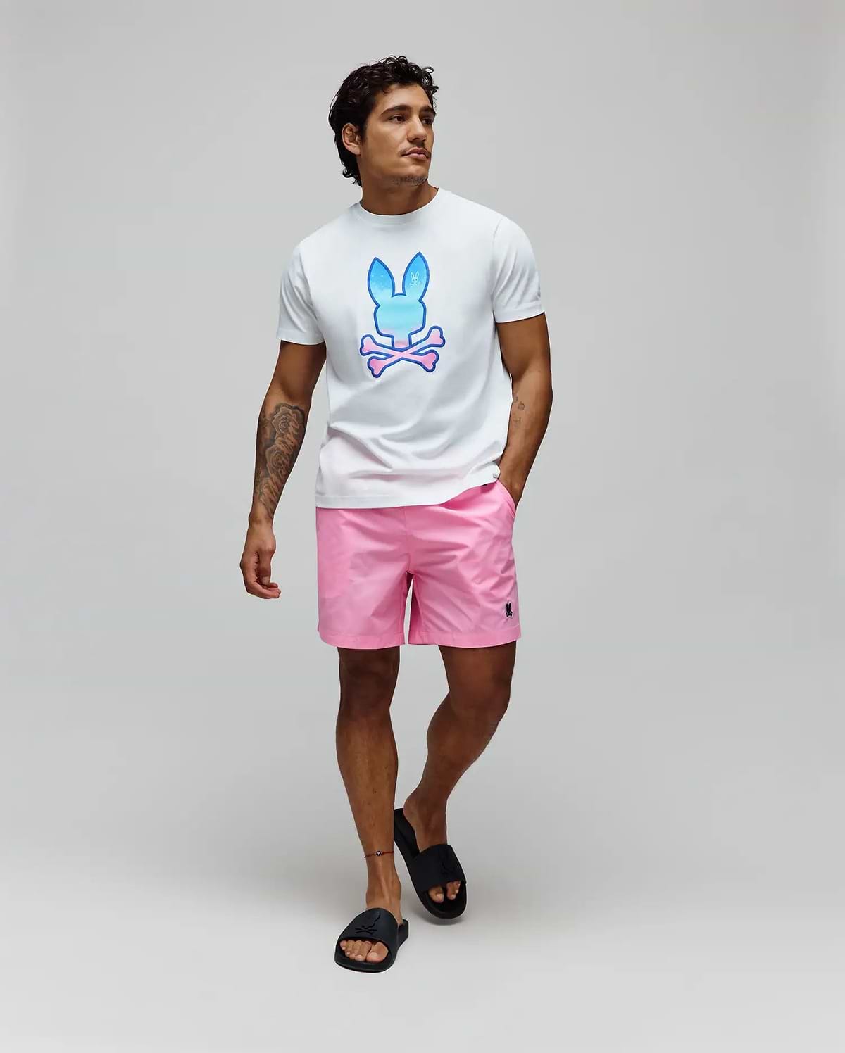 White jersey t-shirt with a large blue and pink gradient Psycho Bunny skull logo on the chest, paired with Pink Mens Samos Hydrochromic Printed Swim Trunk featuring a small black embroidered bunny logo on the left leg.
