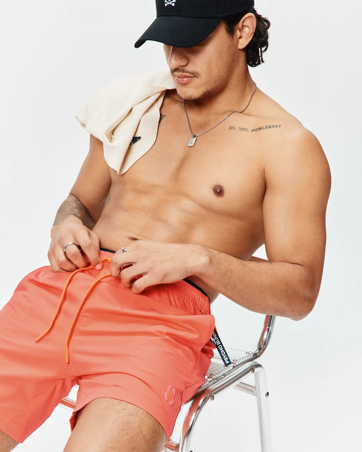 Mens Samos Hydrochromic Printed Swim Trunk in coral with orange drawstrings, a small embroidered Psycho Bunny logo, and branded waistband. Also, a black cap with a white Psycho Bunny skull logo and a cream textured knit towel.