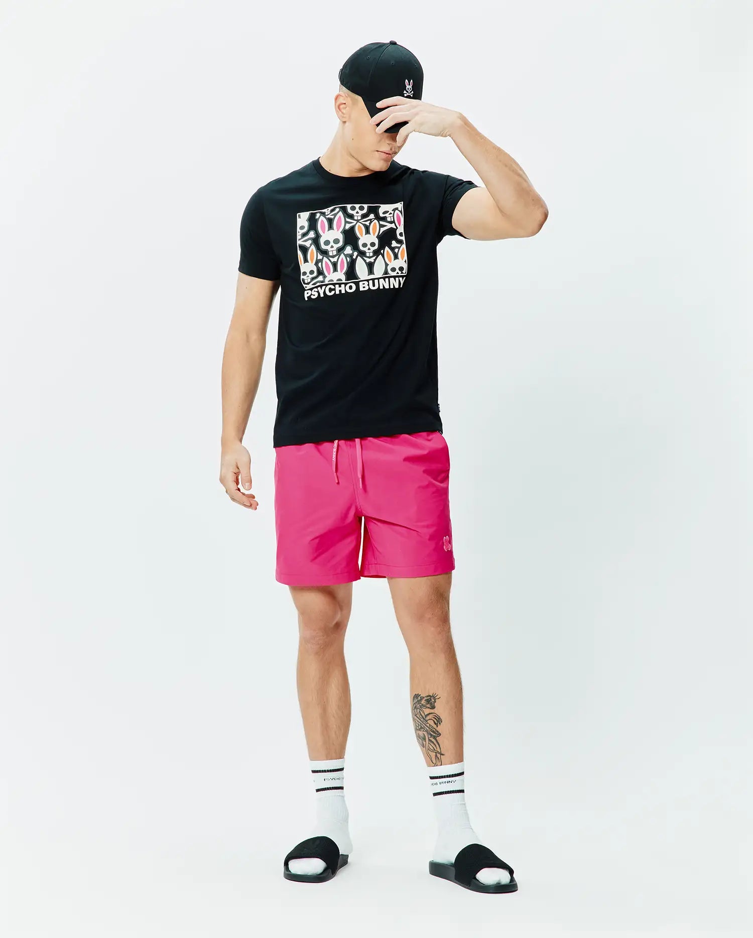 Black classic fit jersey t-shirt with large colorful Psycho Bunny skull graphic, bright pink Mens Samos Hydrochromic Printed Swim Trunk 6 with small embroidered logo, and black cap with pink embroidered Psycho Bunny logo. (6)