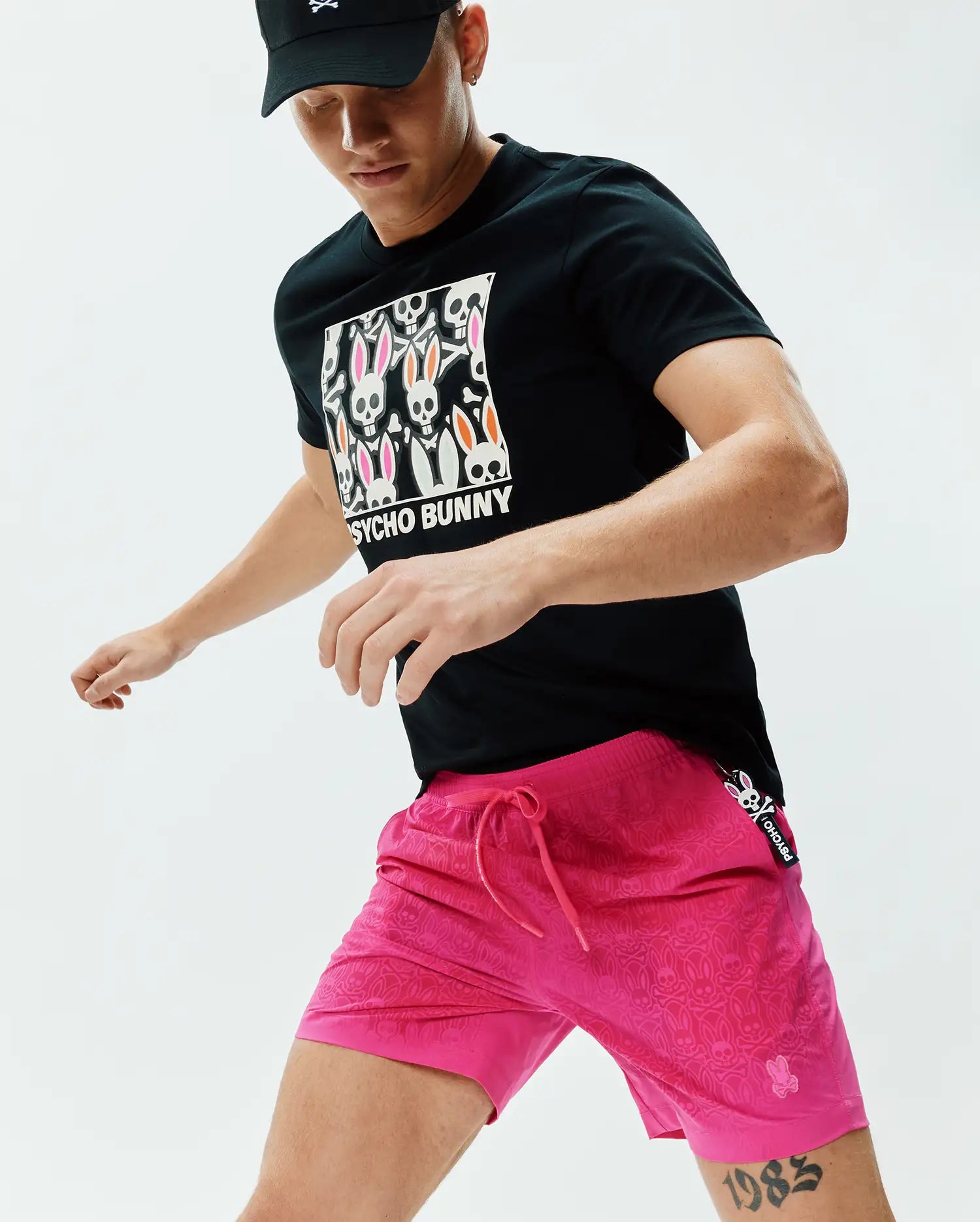 Black classic fit jersey t-shirt with a large chest graphic of colorful Psycho Bunny skull logos and text, paired with bright pink Mens Samos Hydrochromic Printed Swim Trunk 6 featuring an all-over subtle bunny skull pattern and side branding. (5)