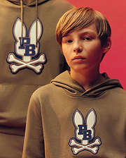 A young boy wears the Psycho Bunny KIDS WADE CORD PATCH HOODIE (KT0500031), featuring the brand’s bunny and crossbones logo, as he stands before an adult in a matching pullover against a reddish backdrop.