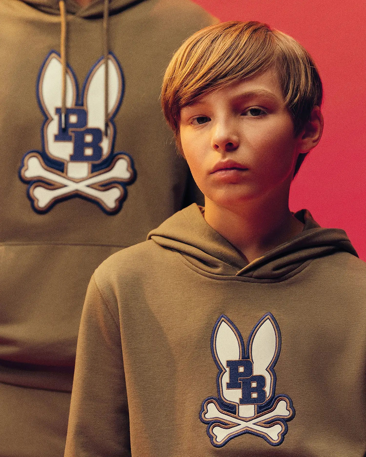 A young boy wears the Psycho Bunny KIDS WADE CORD PATCH HOODIE (KT0500031), featuring the brand’s bunny and crossbones logo, as he stands before an adult in a matching pullover against a reddish backdrop.