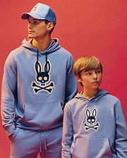 An adult and child wear matching Psycho Bunny MENS RYDER EMBROIDERED BUNNY PATCH HOODIES (MT0500029) in light blue, each with an embroidered Bunny patch. The adult styles it with a blue cap against a pinkish-red background for a modern loungewear look.