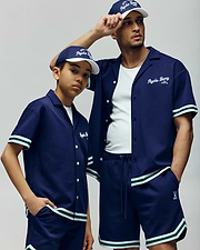 A man and a boy wear matching Psycho Bunny KIDS BASIL TAPED SHORTS (KB0900003) with premium cotton-modal blend, button-up shirts, and baseball caps featuring white and green striped trim as they look confidently into the distance.