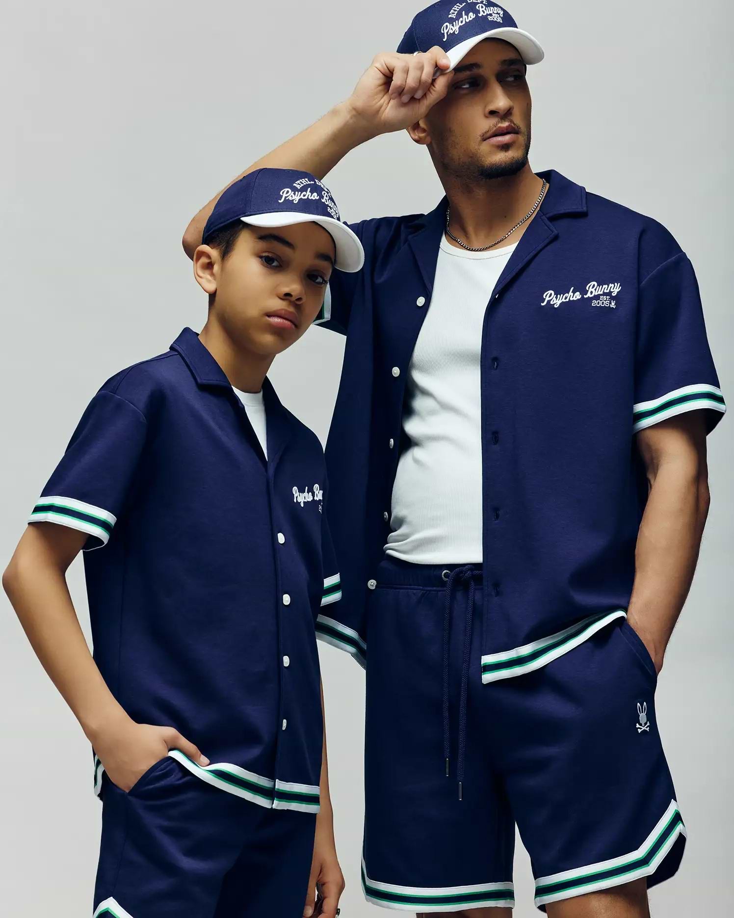 A man and a boy wear matching Psycho Bunny KIDS BASIL TAPED SHORTS (KB0900003) with premium cotton-modal blend, button-up shirts, and baseball caps featuring white and green striped trim as they look confidently into the distance.