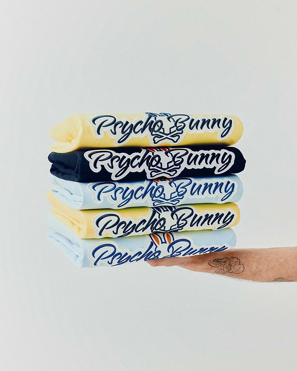 Mens Jaydon Wordmark Graphic Tee stack: yellow, navy, and light blue jersey tees featuring a large embroidered dark blue Psycho Bunny wordmark with white outline and iconic bunny skull logo with red accents on the chest. (5)