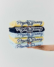 Mens Jaydon Wordmark Graphic Tee stack: light yellow, navy, and light blue jersey tees with large, embroidered Psycho Bunny script logo and iconic bunny skull, some featuring red/orange ear accents. (6)