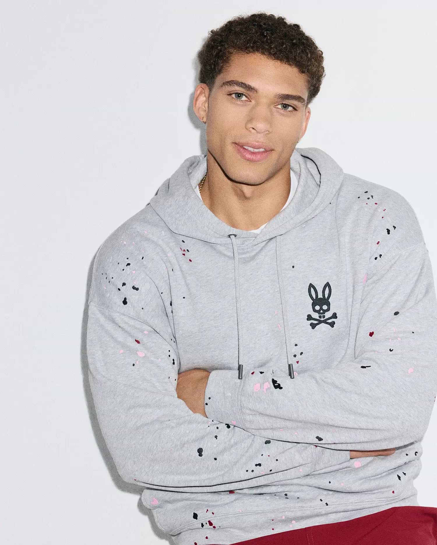 Mens Malcom Splatter Hoodie, a relaxed fit grey french terry hoodie with black, pink, and red splatter paint, featuring an embroidered black Psycho Bunny skull logo on the left chest, and a drawstring hood with a kangaroo pocket.