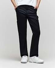 A child stands against a plain background, wearing Psycho Bunny KIDS RUSSELL CARPENTER PANT - KB0600001 in black with cargo pockets, paired with a white shirt and white sneakers. Their hands are in their pockets and their face is not shown.