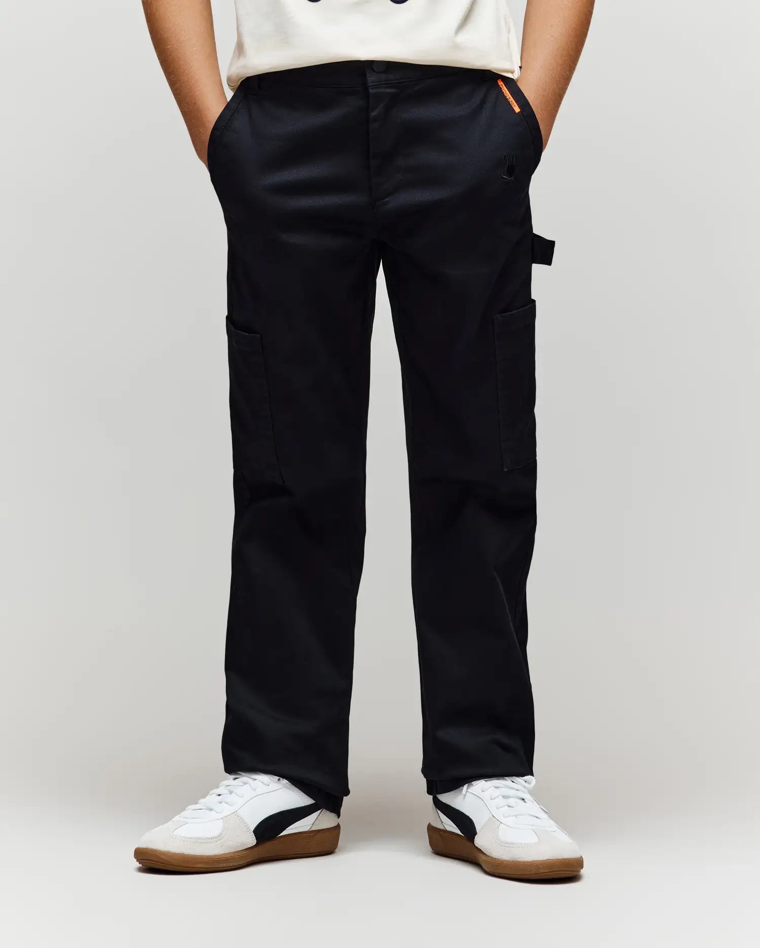 A person models the Psycho Bunny KIDS RUSSELL CARPENTER PANT (KB0600001) with cargo pockets and white sneakers, standing with hands in pockets against a plain light background.