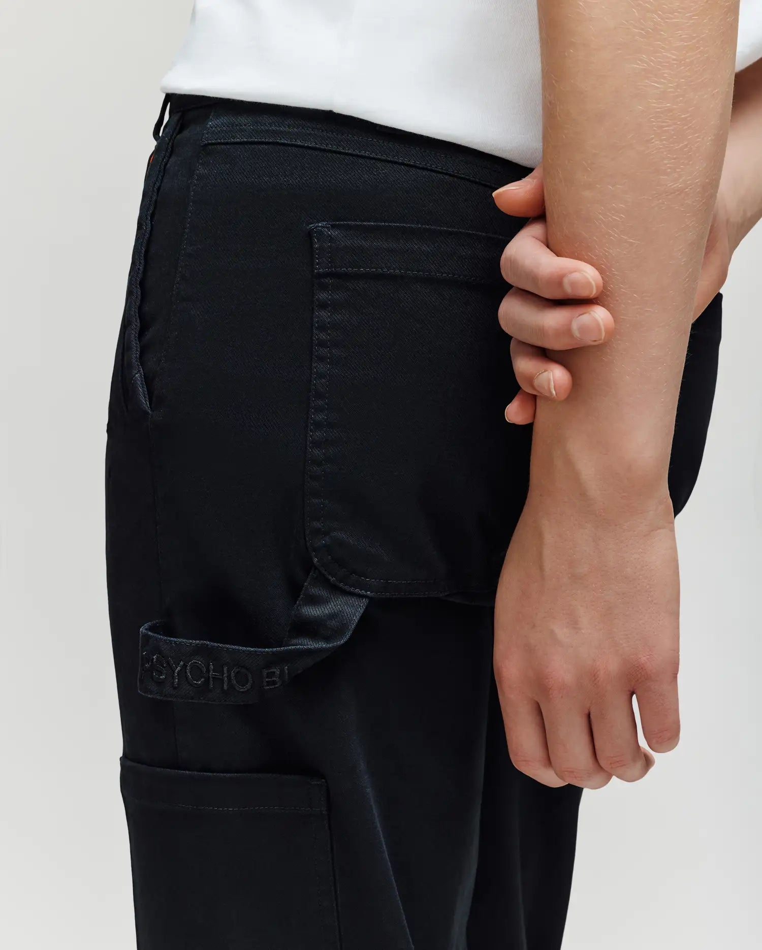 A person grips their opposite arm behind their back, wearing Psycho Bunny KIDS RUSSELL CARPENTER PANT (KB0600001) in a regular fit with visible cargo pockets and a white shirt. The cropped image highlights the waist and hand.