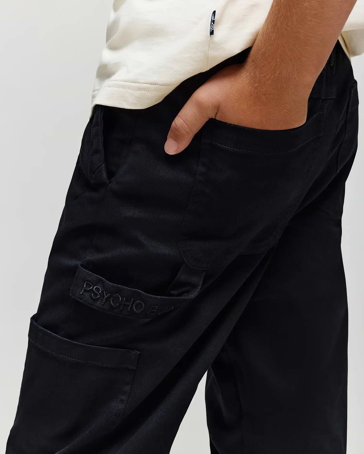A person wears a white shirt and black Psycho Bunny KIDS RUSSELL CARPENTER PANT (KB0600001) with cargo pockets, one hand in their pocket, and a visible "PSYCHO" logo embroidered near the side pocket.