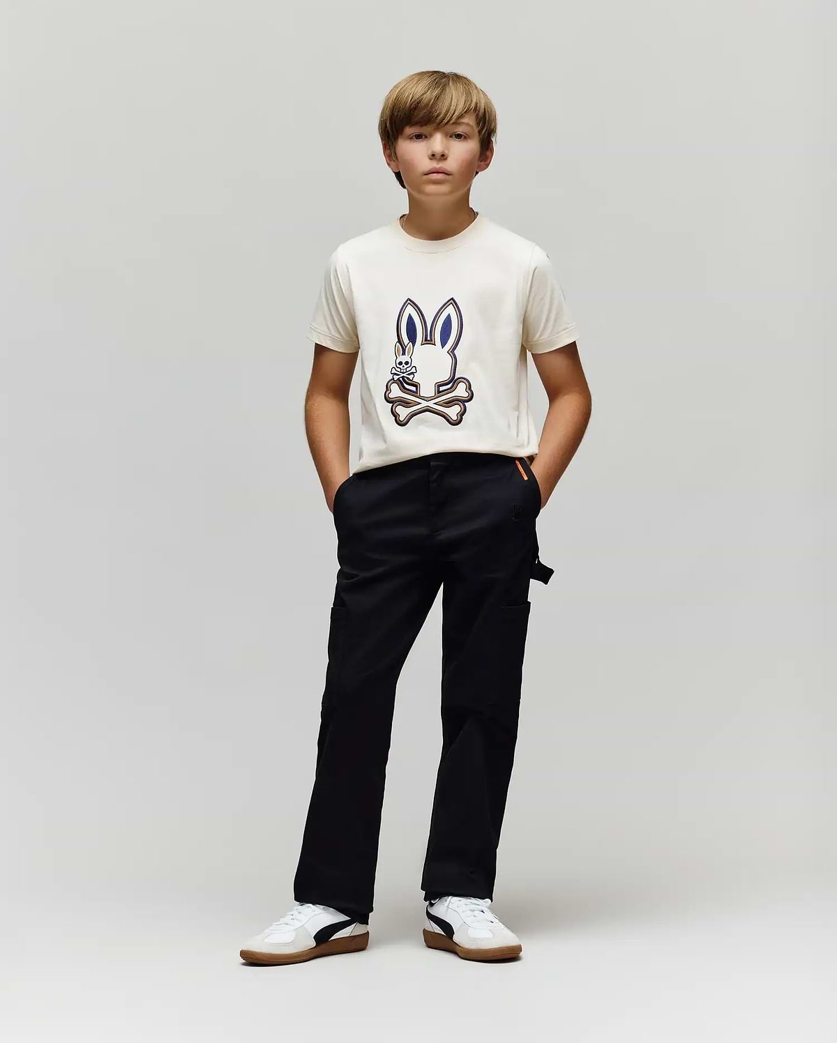 A boy with light brown hair stands against a gray background, wearing a white T-shirt with a cartoon bunny graphic, Psycho Bunny KIDS RUSSELL CARPENTER PANT - KB0600001, and white sneakers. His hands are in his pockets as he looks ahead.