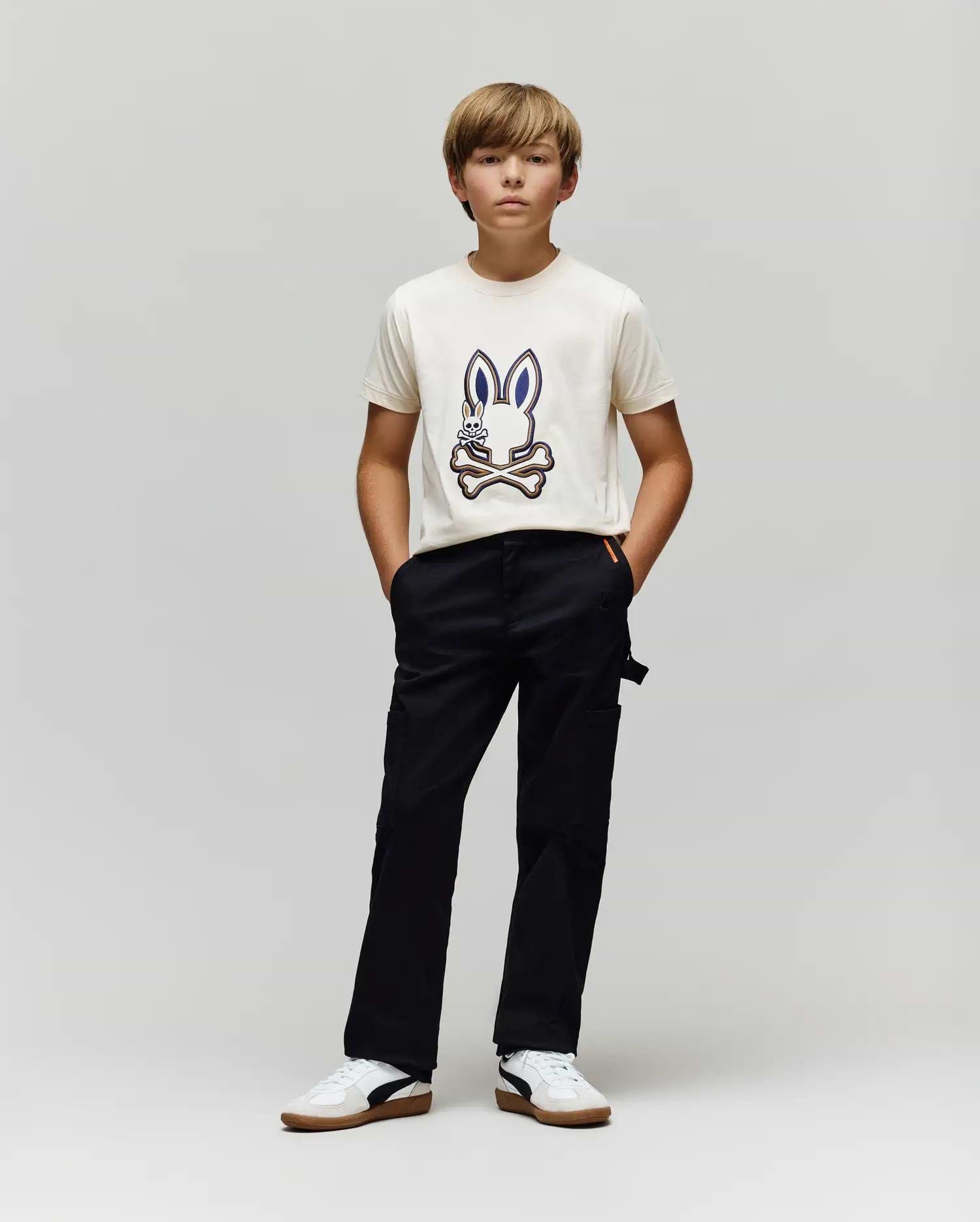 A boy with light brown hair stands against a gray background, wearing a white T-shirt with a cartoon bunny graphic, Psycho Bunny KIDS RUSSELL CARPENTER PANT - KB0600001, and white sneakers. His hands are in his pockets as he looks ahead.