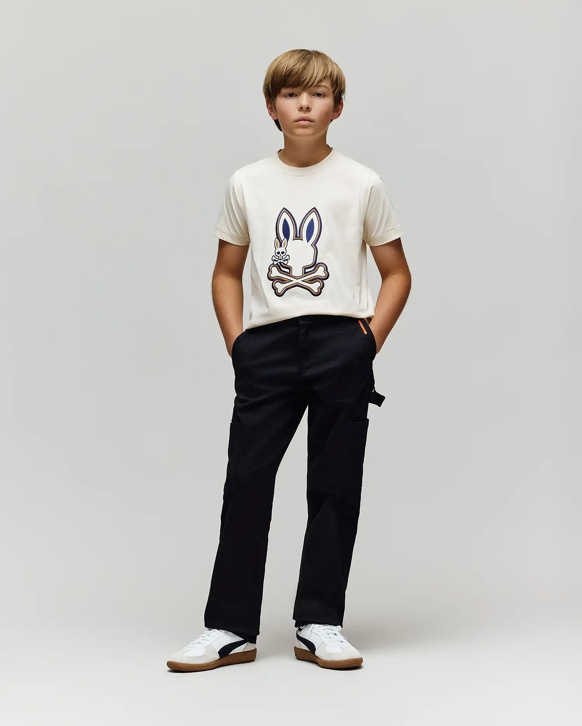 A boy with light brown hair stands against a gray background, wearing a white T-shirt with a cartoon bunny graphic, Psycho Bunny KIDS RUSSELL CARPENTER PANT - KB0600001, and white sneakers. His hands are in his pockets as he looks ahead.