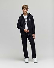 A teenage boy stands against a plain background wearing a black hoodie, Psycho Bunny KIDS RUSSELL CARPENTER PANT - KB0600001, white sneakers, and a white T-shirt with a large bunny graphic.