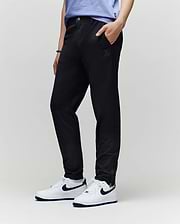 A person in Psycho Bunny KIDS MATHIEU COMMUTER PANT (KB0600002), a light blue shirt, and white Nike sneakers with a dark blue swoosh stands with one hand in their pocket against a plain light background.