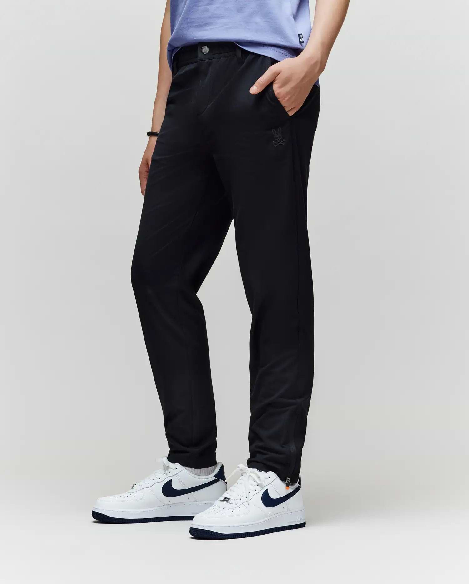 A person in Psycho Bunny KIDS MATHIEU COMMUTER PANT (KB0600002), a light blue shirt, and white Nike sneakers with a dark blue swoosh stands with one hand in their pocket against a plain light background.