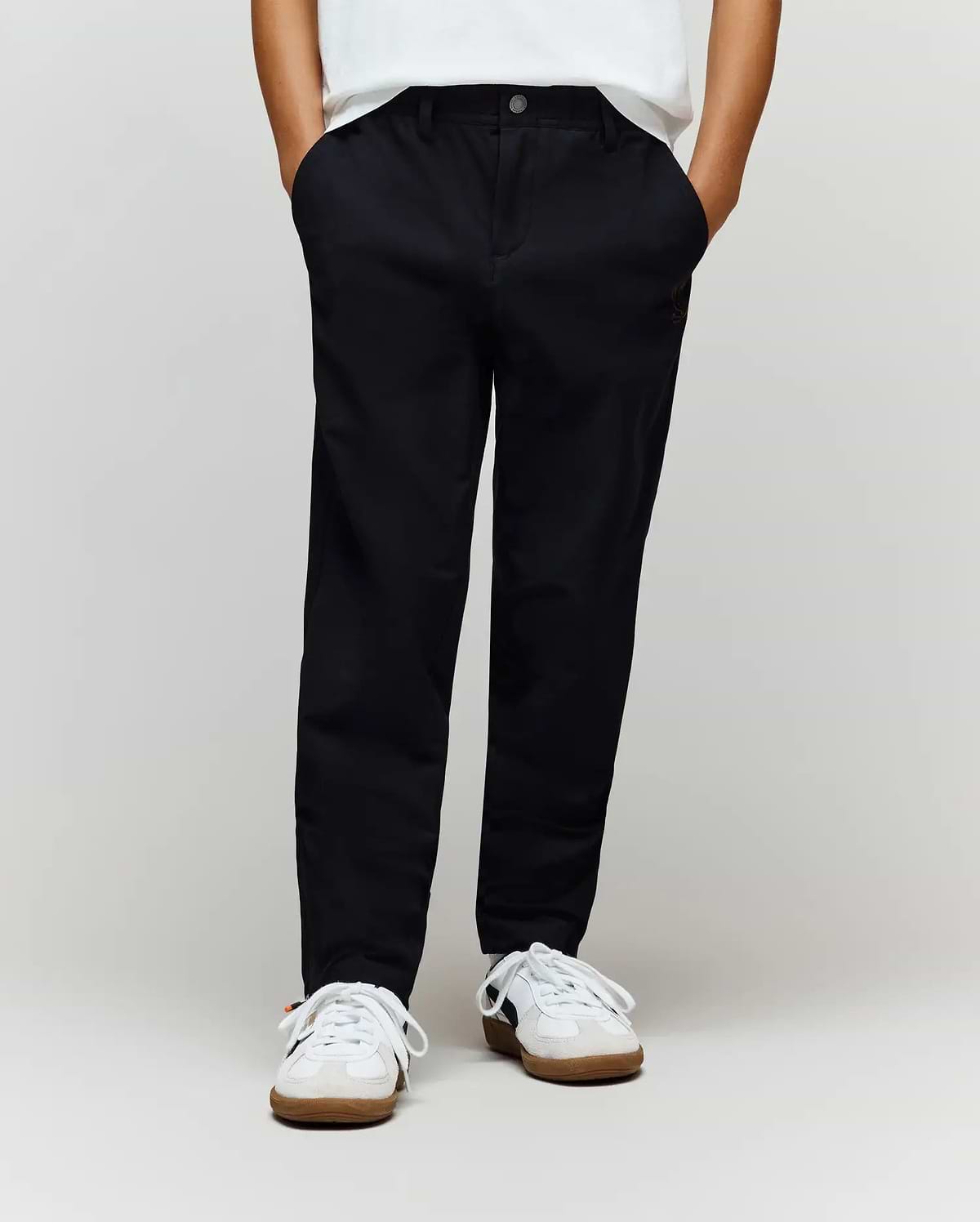 A child wears Psycho Bunny’s KIDS MATHIEU COMMUTER PANT (KB0600002) in classic black with subtle Bunny embroidery, paired with white sneakers, hands in pockets. Only the lower half is shown against a plain light background.