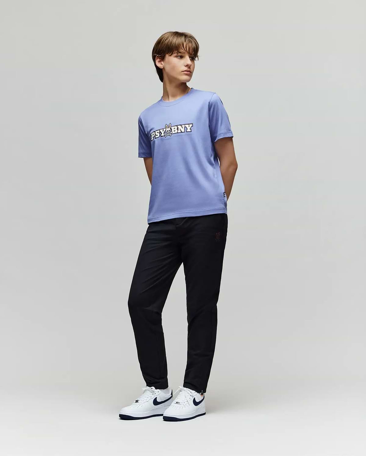 A young person stands against a plain background in Psycho Bunny's KIDS MATHIEU COMMUTER PANT (KB0600002), a lavender "PSYBNY" T-shirt, and white Nike sneakers with black swooshes, glancing sideways with hands behind their back.