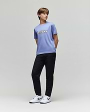 A young person stands against a plain background in Psycho Bunny's KIDS MATHIEU COMMUTER PANT (KB0600002), a lavender "PSYBNY" T-shirt, and white Nike sneakers with black swooshes, glancing sideways with hands behind their back.