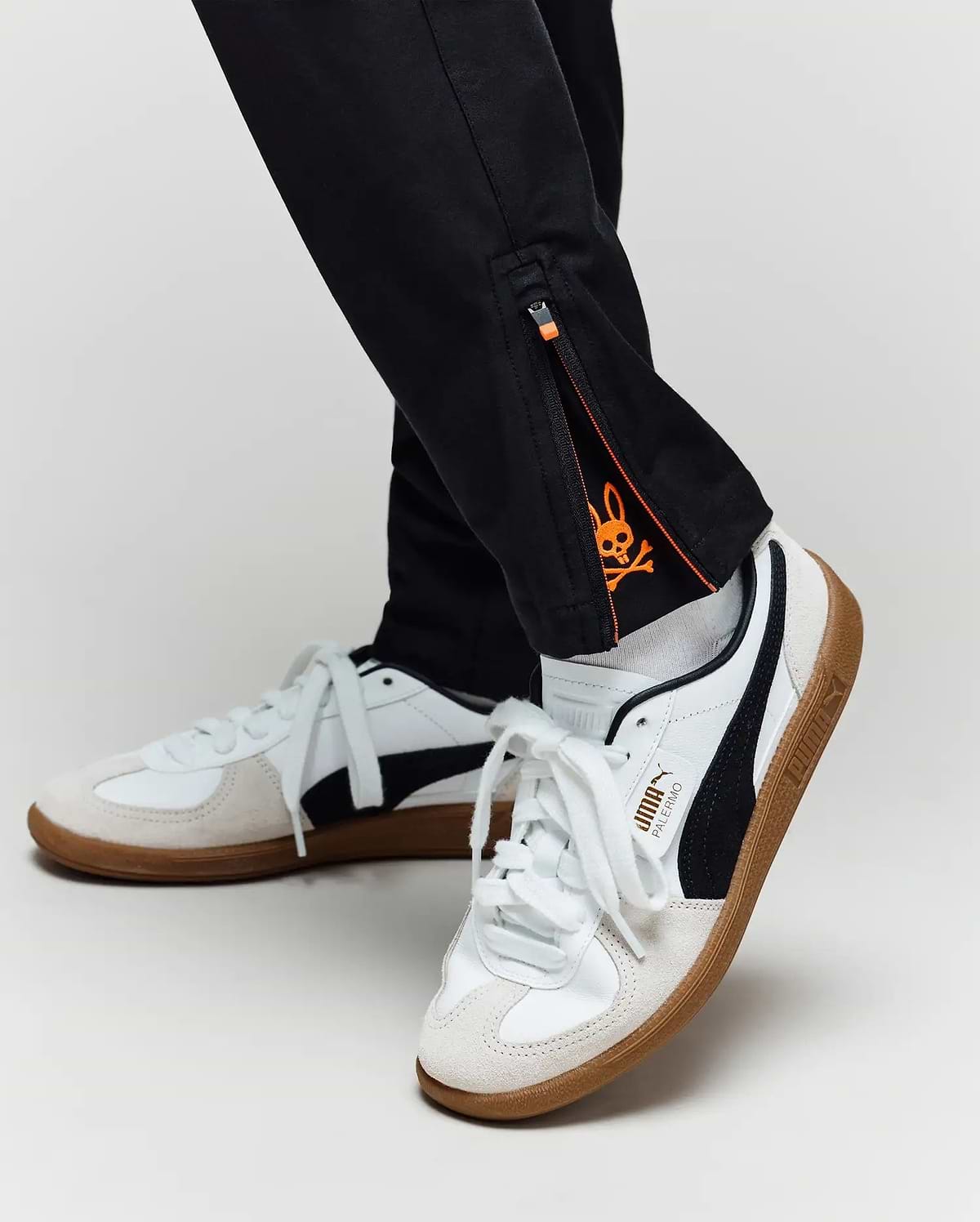 A close-up of a person in Psycho Bunny KIDS MATHIEU COMMUTER PANT (KB0600002) featuring an orange skull graphic inside the ankle zipper, paired with white and black Puma sneakers with gum soles.