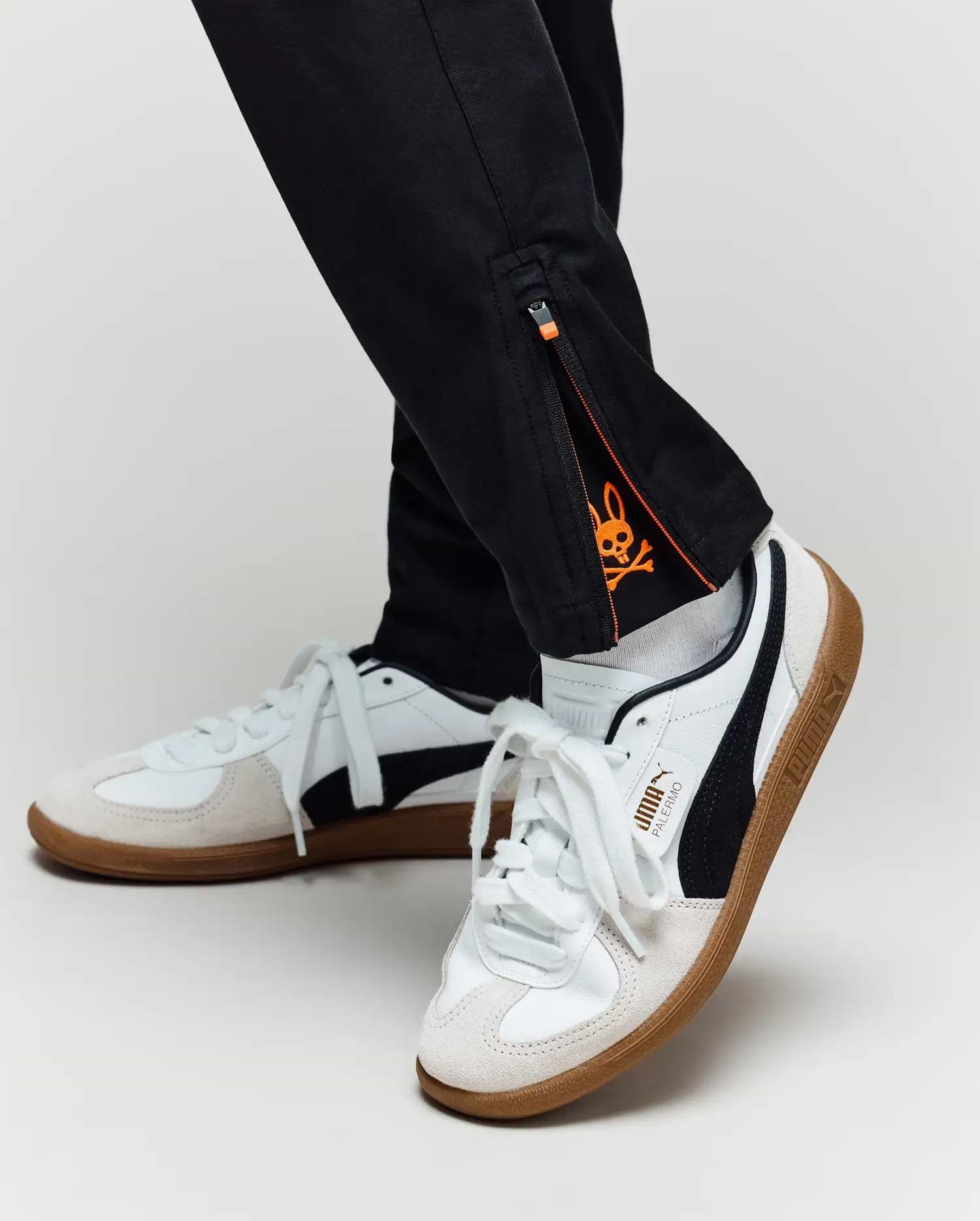 A close-up of a person in Psycho Bunny KIDS MATHIEU COMMUTER PANT (KB0600002) featuring an orange skull graphic inside the ankle zipper, paired with white and black Puma sneakers with gum soles.