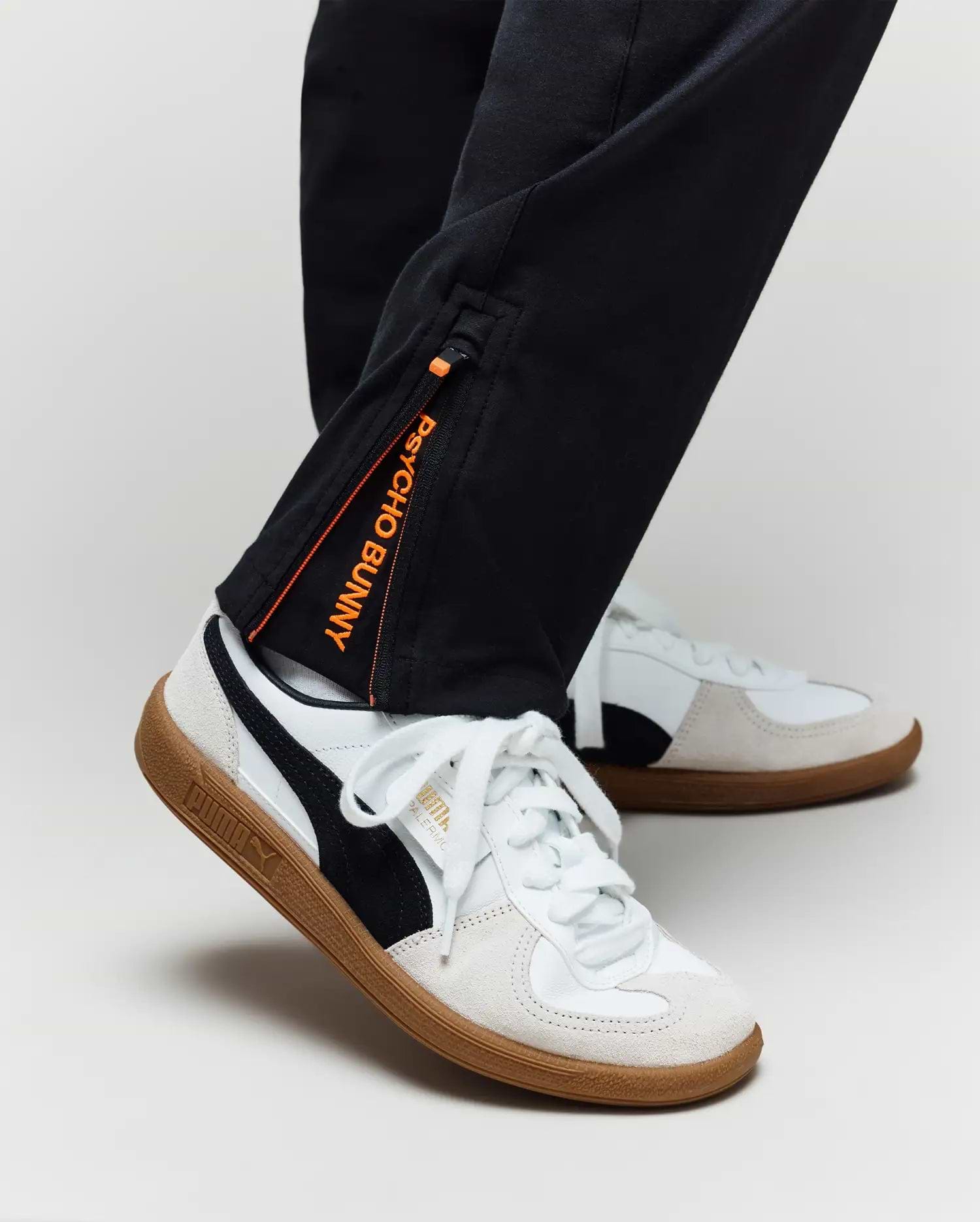 A person wearing Psycho Bunny KIDS MATHIEU COMMUTER PANT (KB0600002), classic fit black pants with Bunny embroidery and an orange tag, paired with white Puma sneakers with black stripes and gum soles, shown from the ankle down.