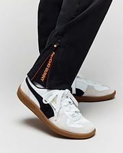 A person wearing Psycho Bunny KIDS MATHIEU COMMUTER PANT (KB0600002), classic fit black pants with Bunny embroidery and an orange tag, paired with white Puma sneakers with black stripes and gum soles, shown from the ankle down.