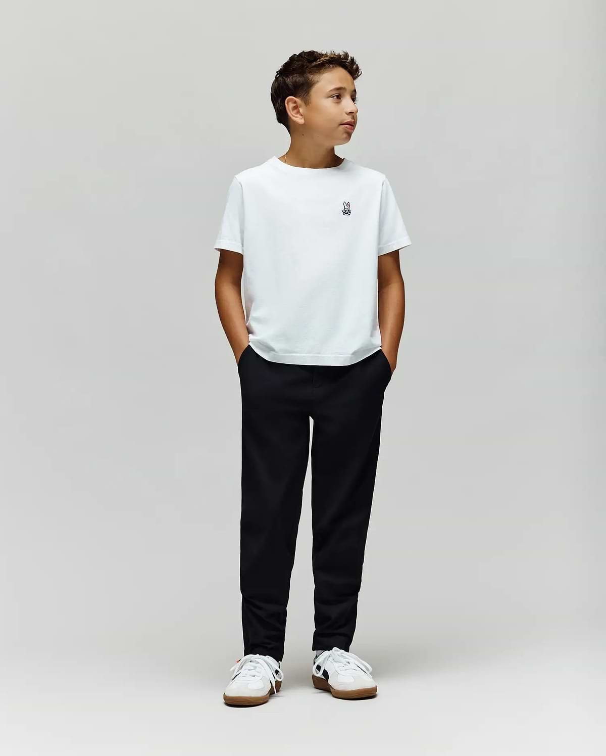 A boy stands against a plain light gray background, wearing a white t-shirt, Psycho Bunny KIDS MATHIEU COMMUTER PANT (KB0600002), and white sneakers. His hands are in his pockets, looking to the side.