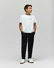 A boy stands against a plain light gray background, wearing a white t-shirt, Psycho Bunny KIDS MATHIEU COMMUTER PANT (KB0600002), and white sneakers. His hands are in his pockets, looking to the side.